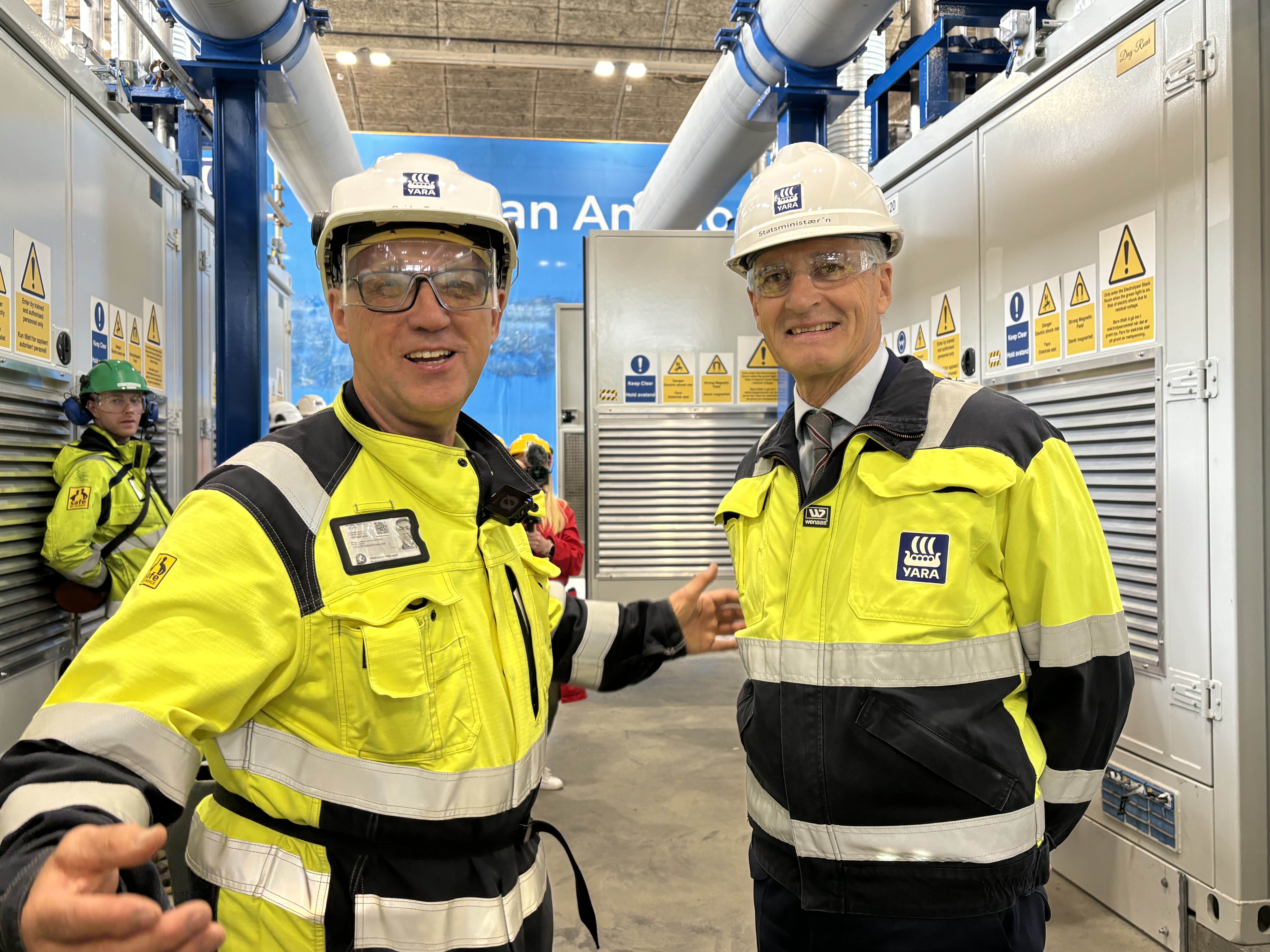 to menn i gult og blått verneutstyr, hjelm og briller, står i nytt hydrogenfabrikkanlegg. Åpningsseremoni.