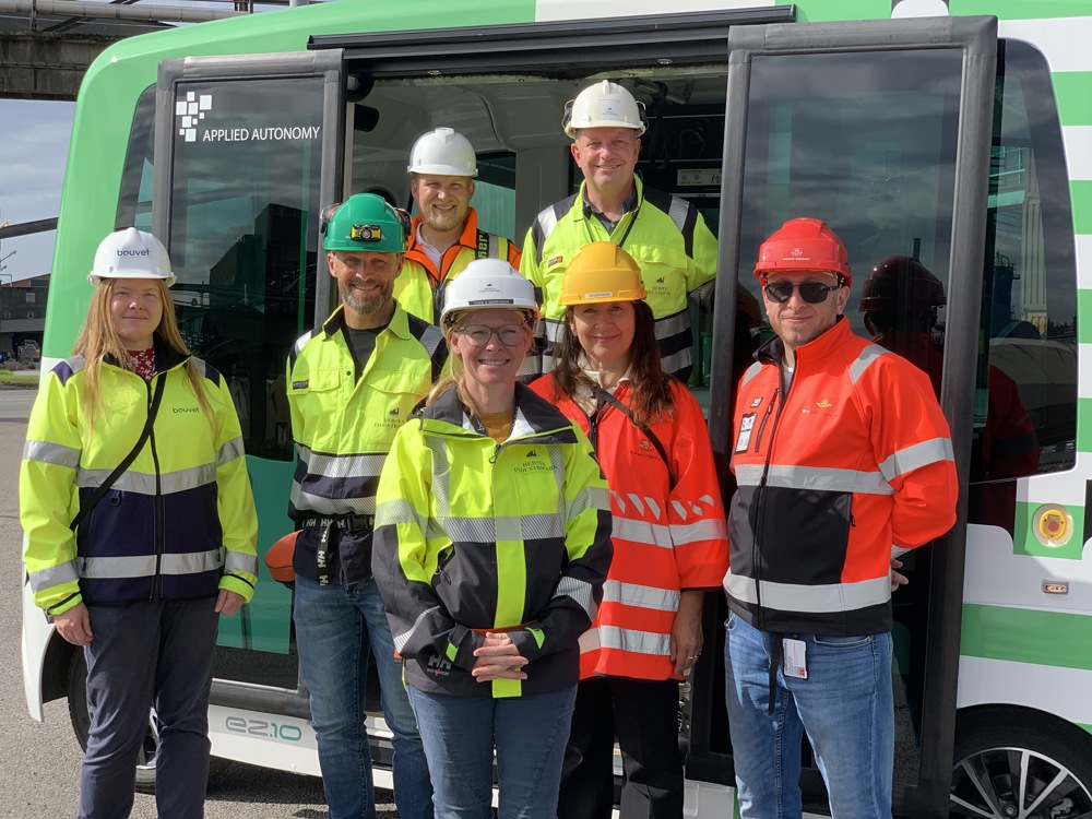 syv personer, gruppebilde, poserer, i og rundt døråpningen på en hvit og grønn autonom minibuss.