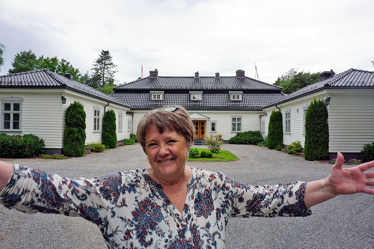 kvinne står med armene ut, ønsker velkommen, til stort hvitt trehus i bakgrunnen. Hun poserer, og smiler. Trehus har hage og stor hovedinngang. Representasjonsbolig.