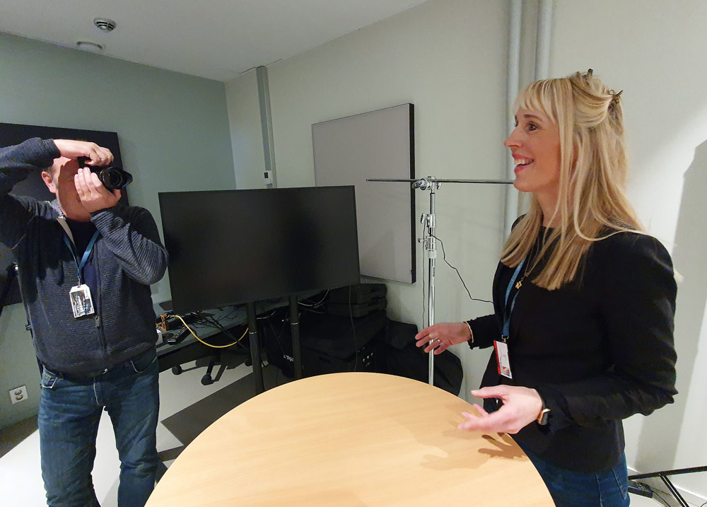 in a studio, woman standing at a high table, presenting, man standing on the other side of table holding a camera. Studio equipment.