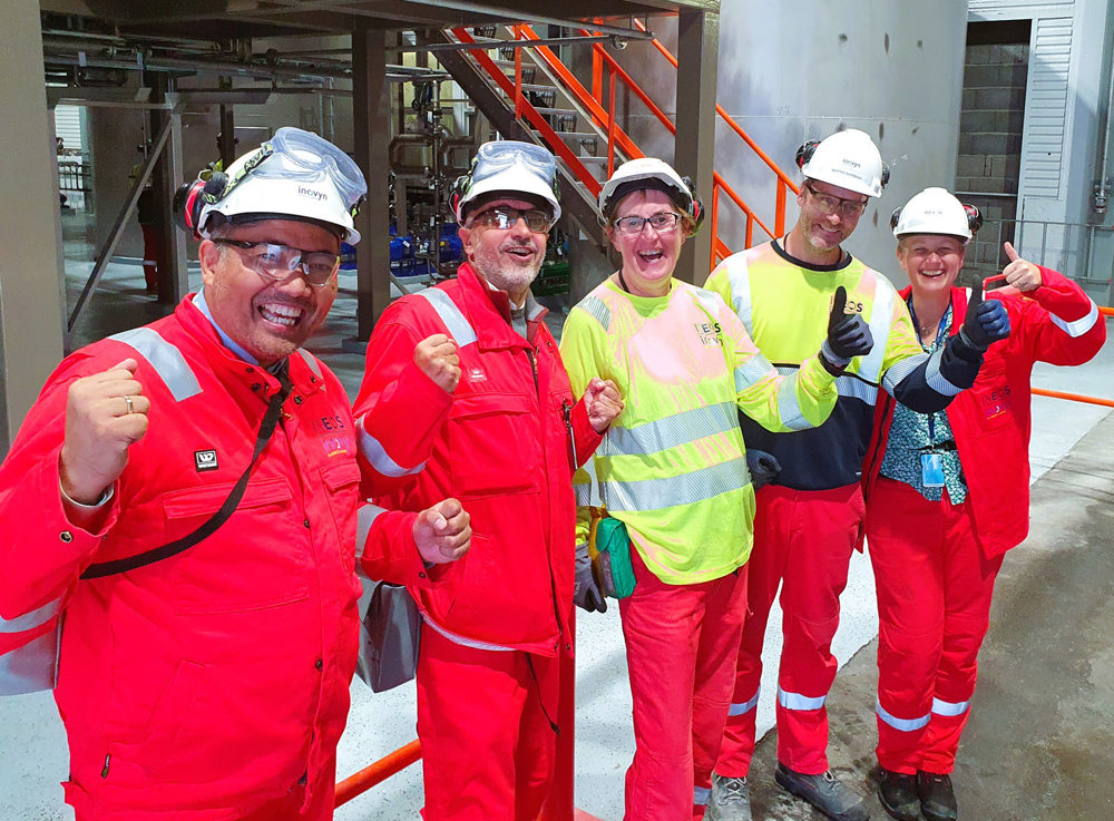 five people standing in line, all smiling, posing, yellow and red work clothes, white helmets, standing in a production hall.