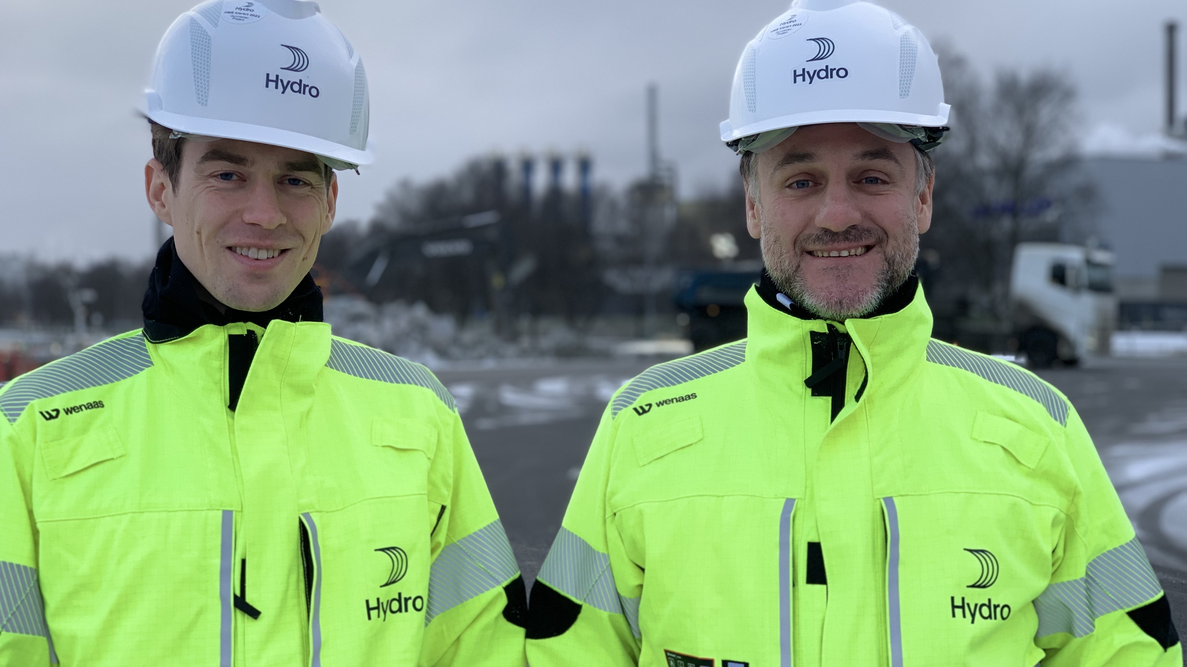 two men, portrait, posing, white helmet, yellow jackets, construction site, large vehicles and escavator in background, fjord
