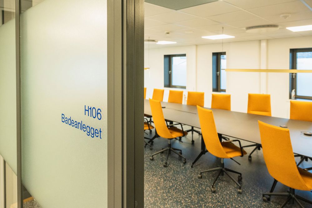 viewing a meeting room with long table and many orange chairs, through a glass door.