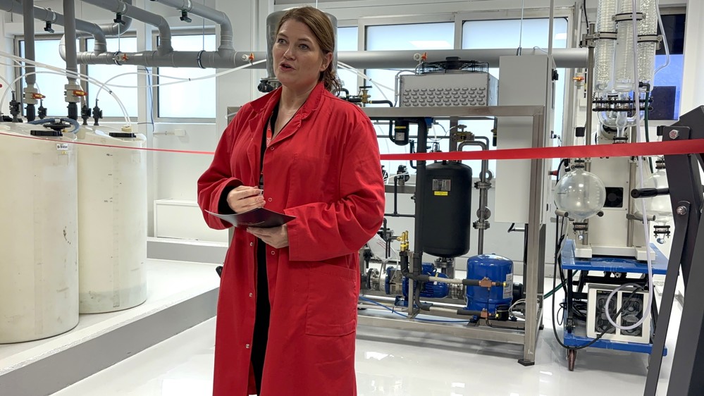 young woman dressed in red visitors coat, speaks. Pilot facility in background.