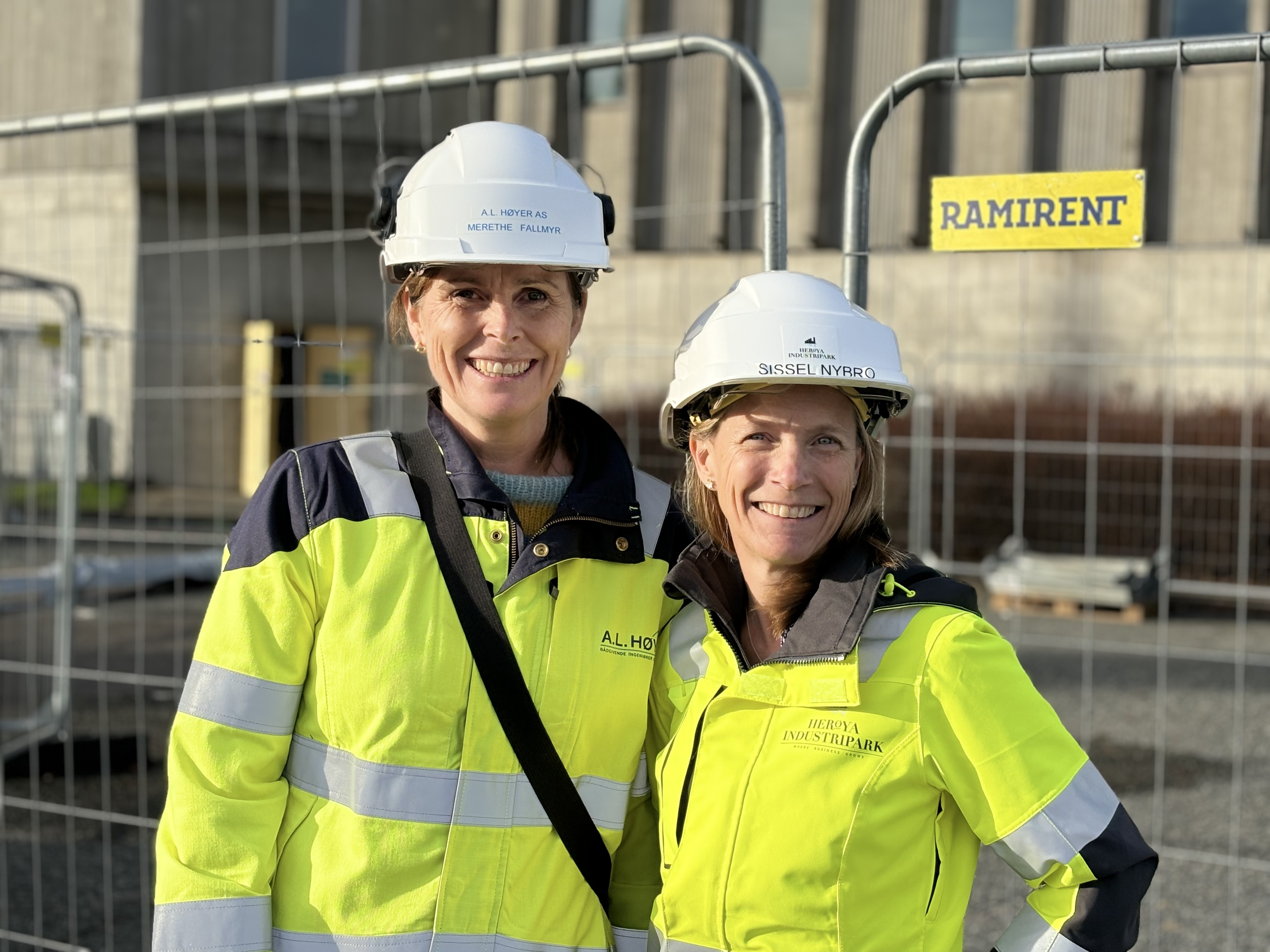 to kvinner i gult arbeidstøy, hvit hjelm, poserer, står foran stålgjerde, byggeplass.