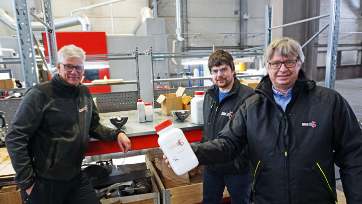 three men posing, standing in a workshop, one of them holding a white plastic container with a red lid in his hand.