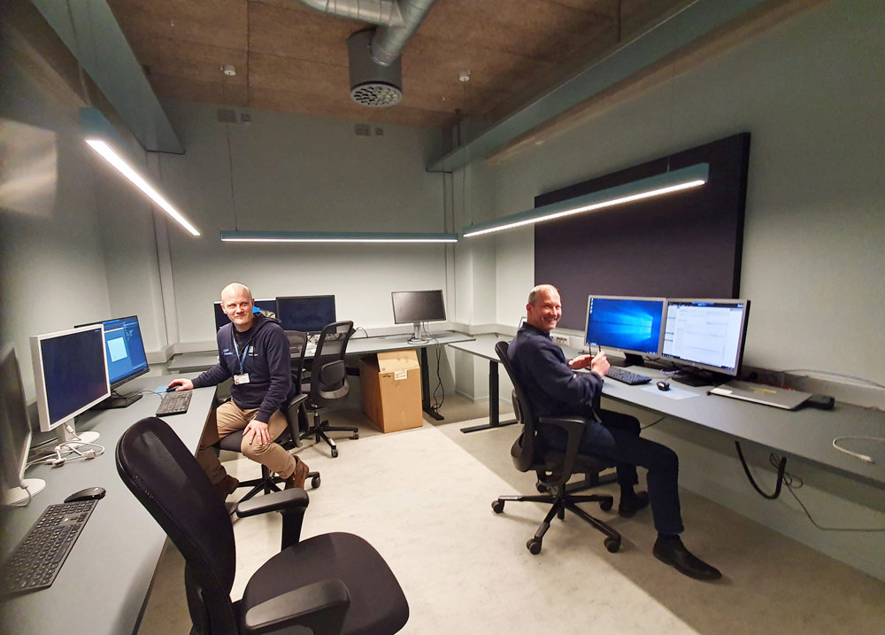to menn sitter ved hver sin pc og monitorer i et stort kontorareal