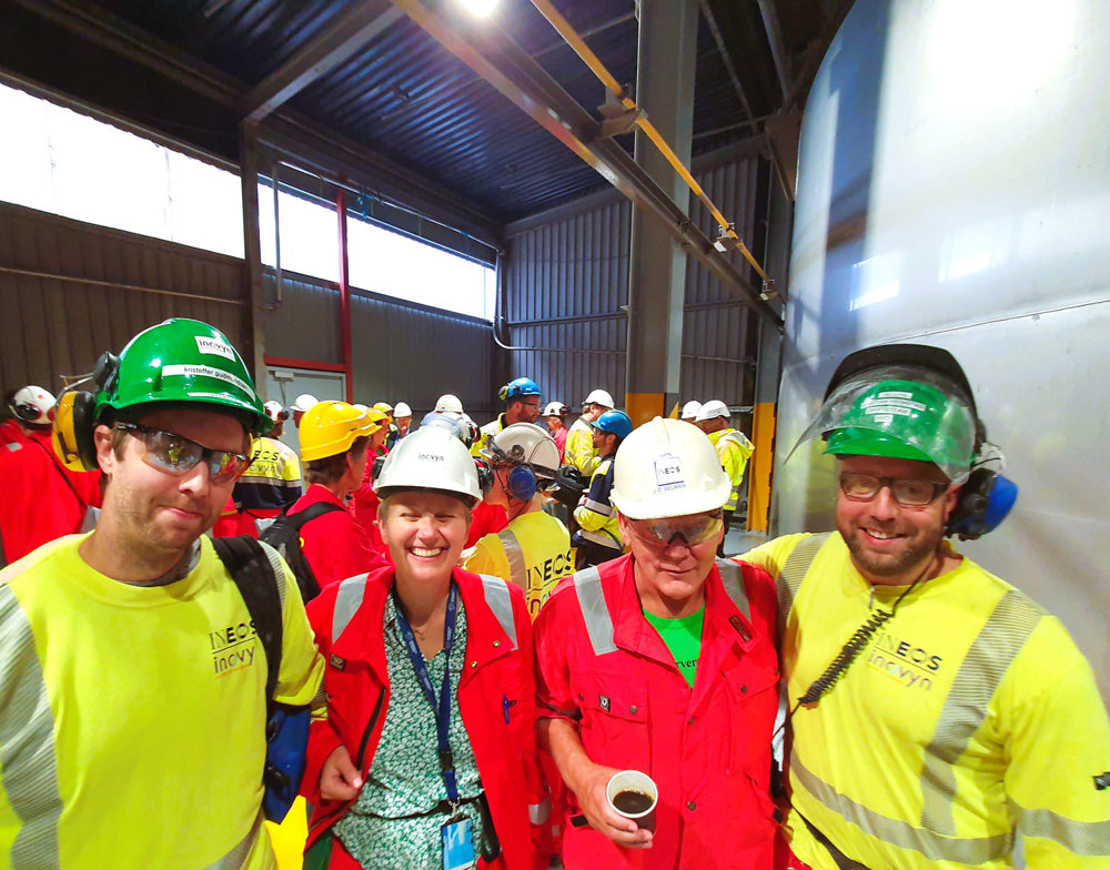 four people standing together, dressed in yellow and red personal protective equipment, posing, smiling, standing in a production hall.