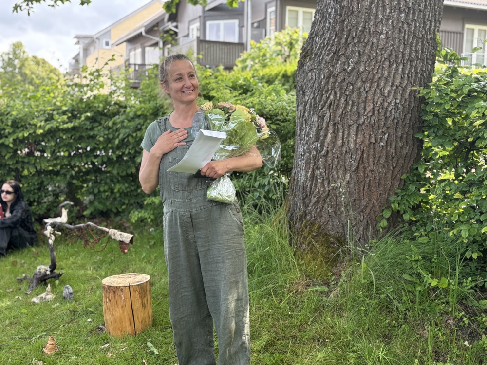 kvinne står ved en tykk trestamme, blomsterbukett i hånden.