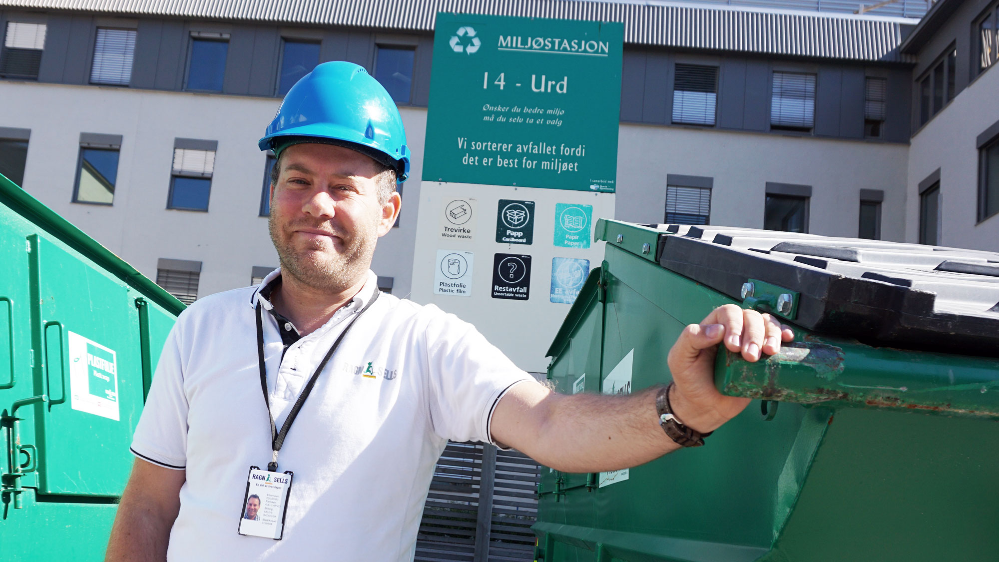 mann i hvit t-skjorte, blå hjelm, poserer, en hånd på grønn avfallscontainer, stort bygg i bakgrunnen