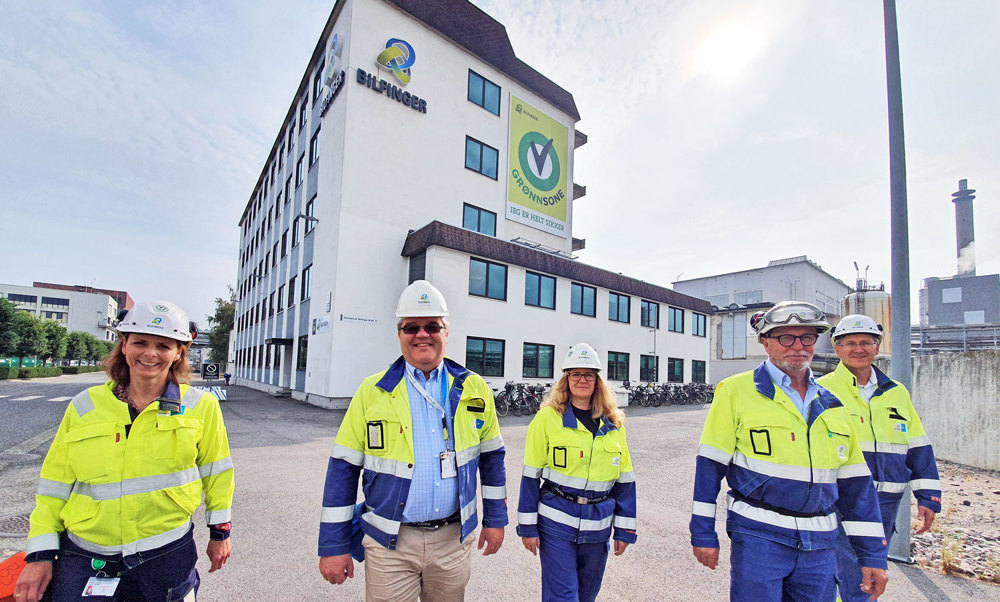 fem personer i arbeidstøy går i gate i industripark. stort bygg med bedriftslogo i bakgrunnen.