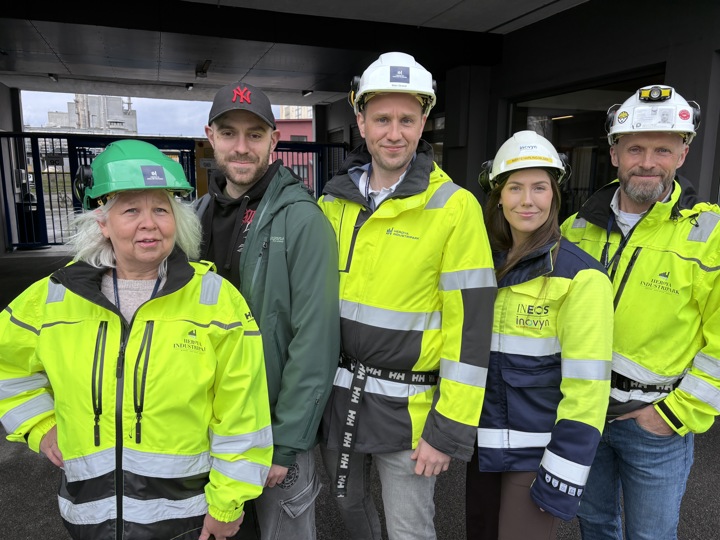 fem personer poserer, står på rekke, ikledd verneutstyr, fabrikkanlegg i bakgrunnen