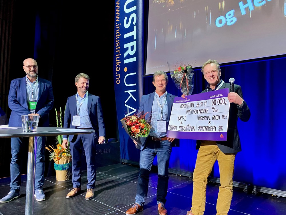 fire personer står på en scene, blomster, stor gavesjekk, høyt bord med vannglass, banner og lerret med presentasjon.