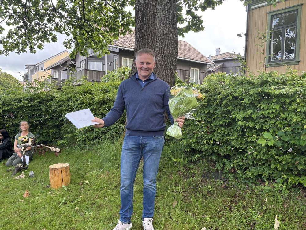 smilende mann, armene ut til siden med blomsterbukett. Grønt parkområde.