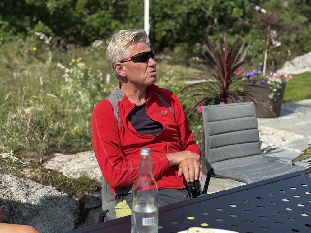 mann i rød jakke, sitter på en stol i hage, solbriller.