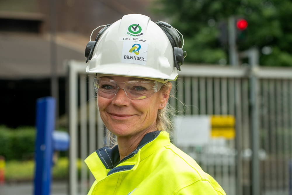 portrett av kvinne, blondt hår, hvit hjelm, gul arbeidsjakke. Industriport i bakgrunnen. Foto: Ole Bjørn Ulsnæs.