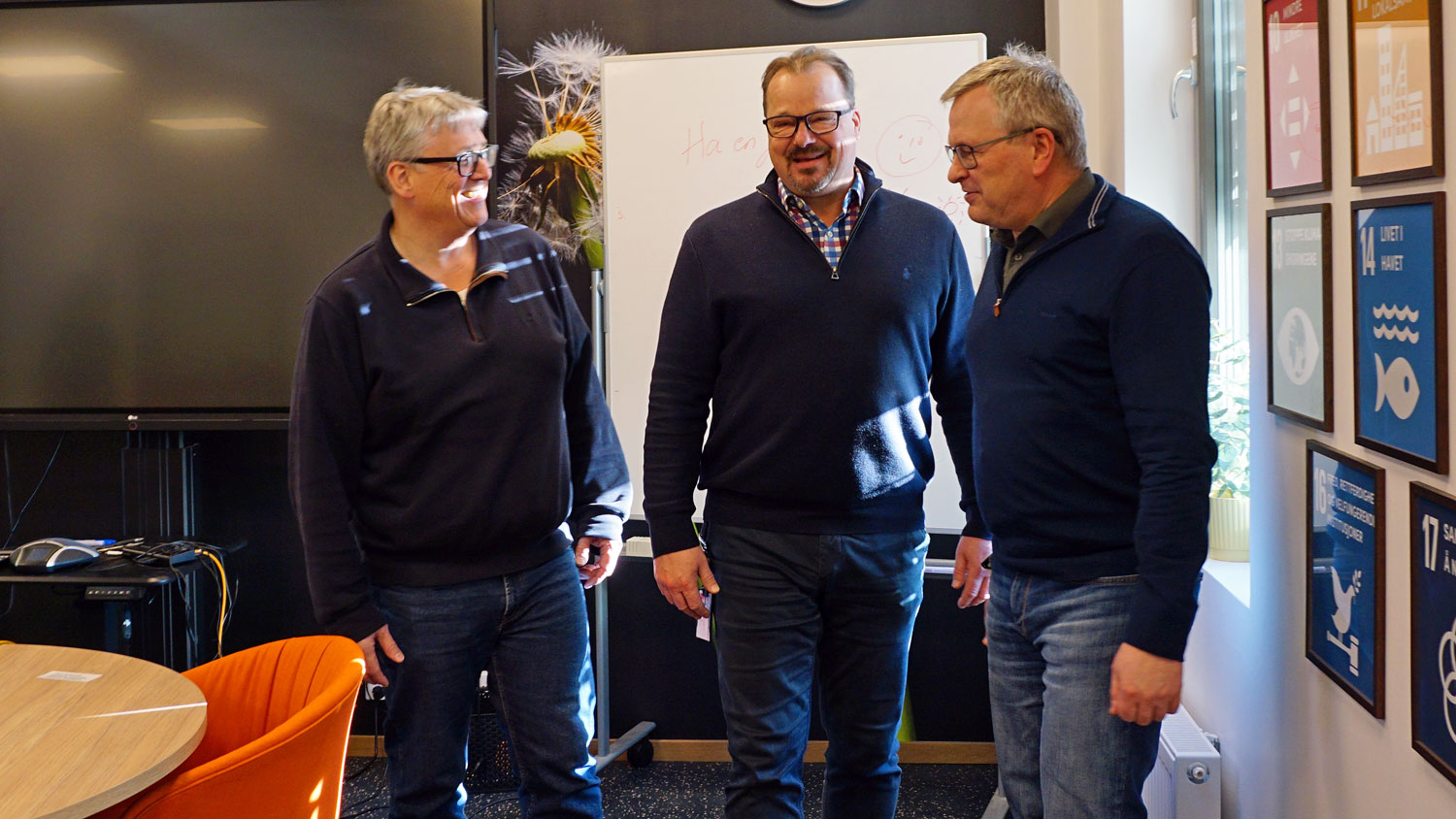 three men standing together, talking, office environment, colourful furniture, monitor on the wall in background