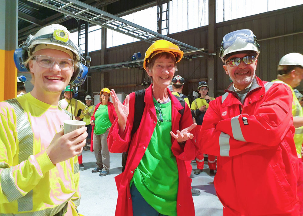 three people standing in a production hall, smiling, posing, all wearing personal protective equipment.