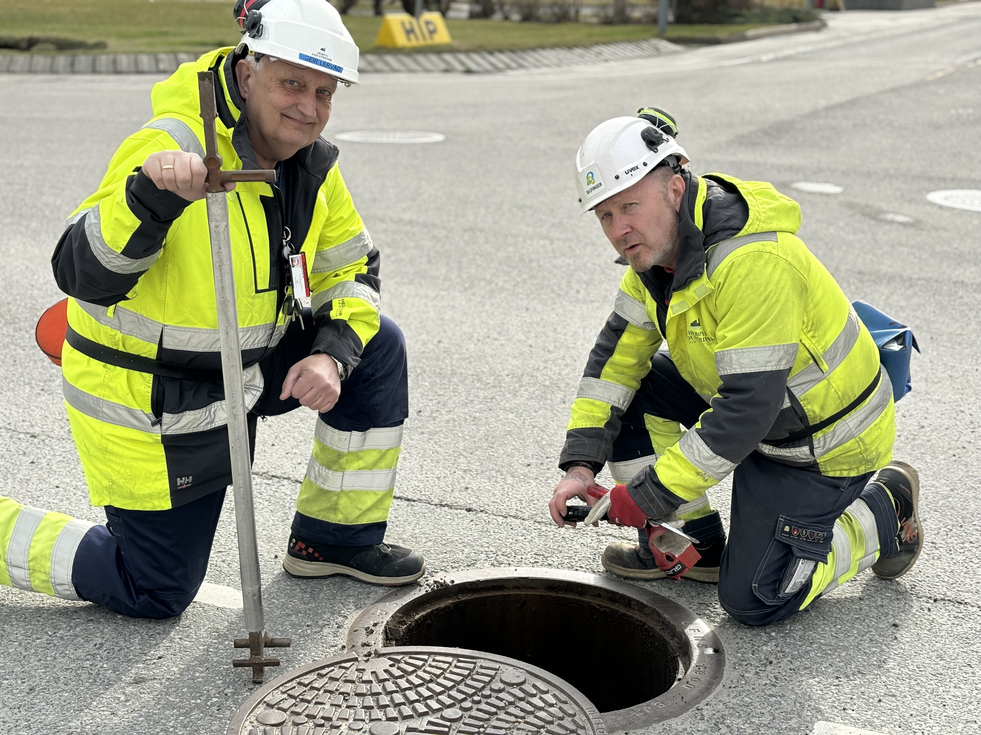Two men are crouching by a manhole in the street, the manhole cover is next to them. One man is holding a crowbar that the manhole cover is opened with, the other is using a camera down into the manhole and taking a picture.