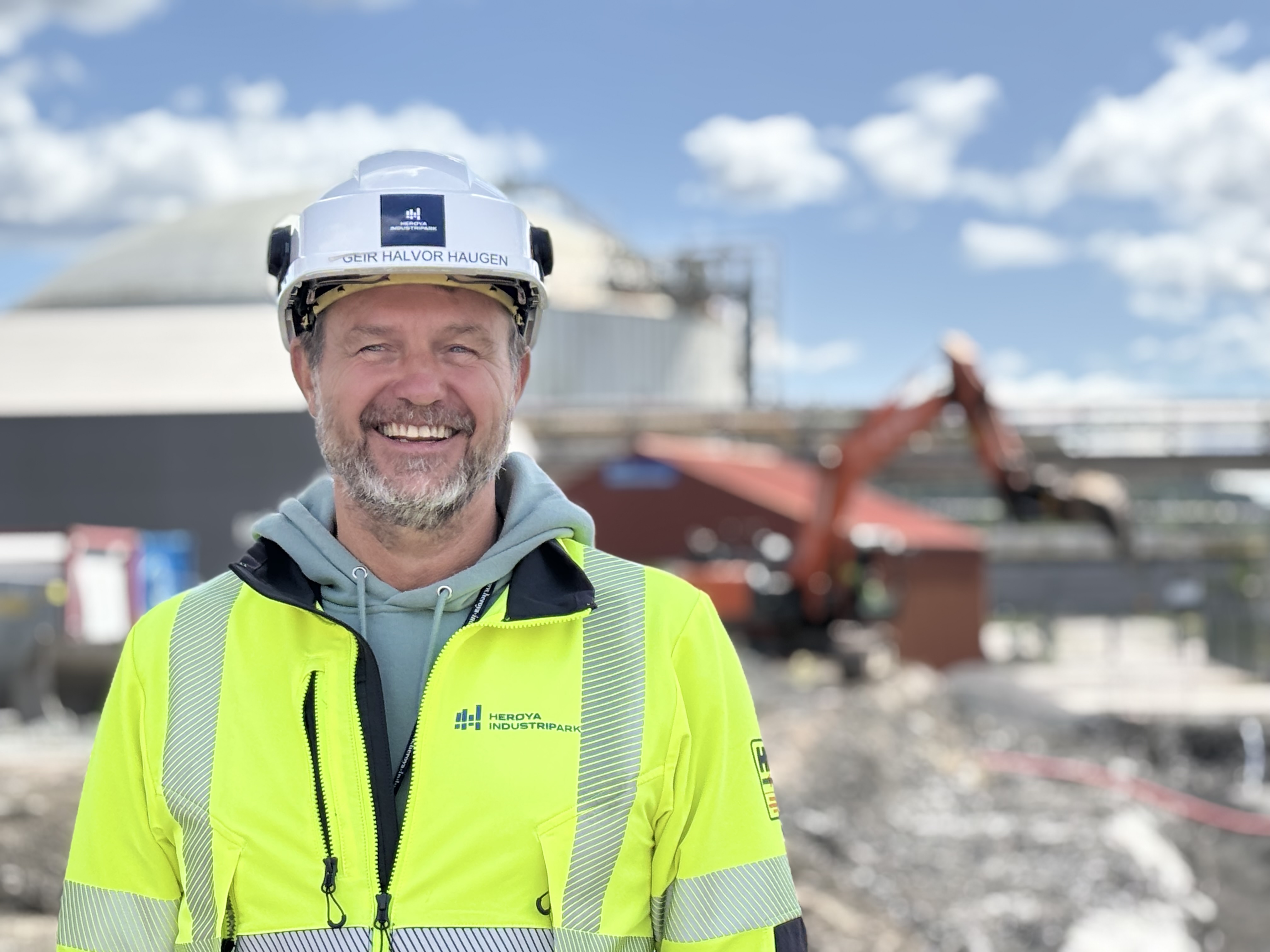 mann i gul jakke og hvit hjelm står ute i anleggsområde industripark