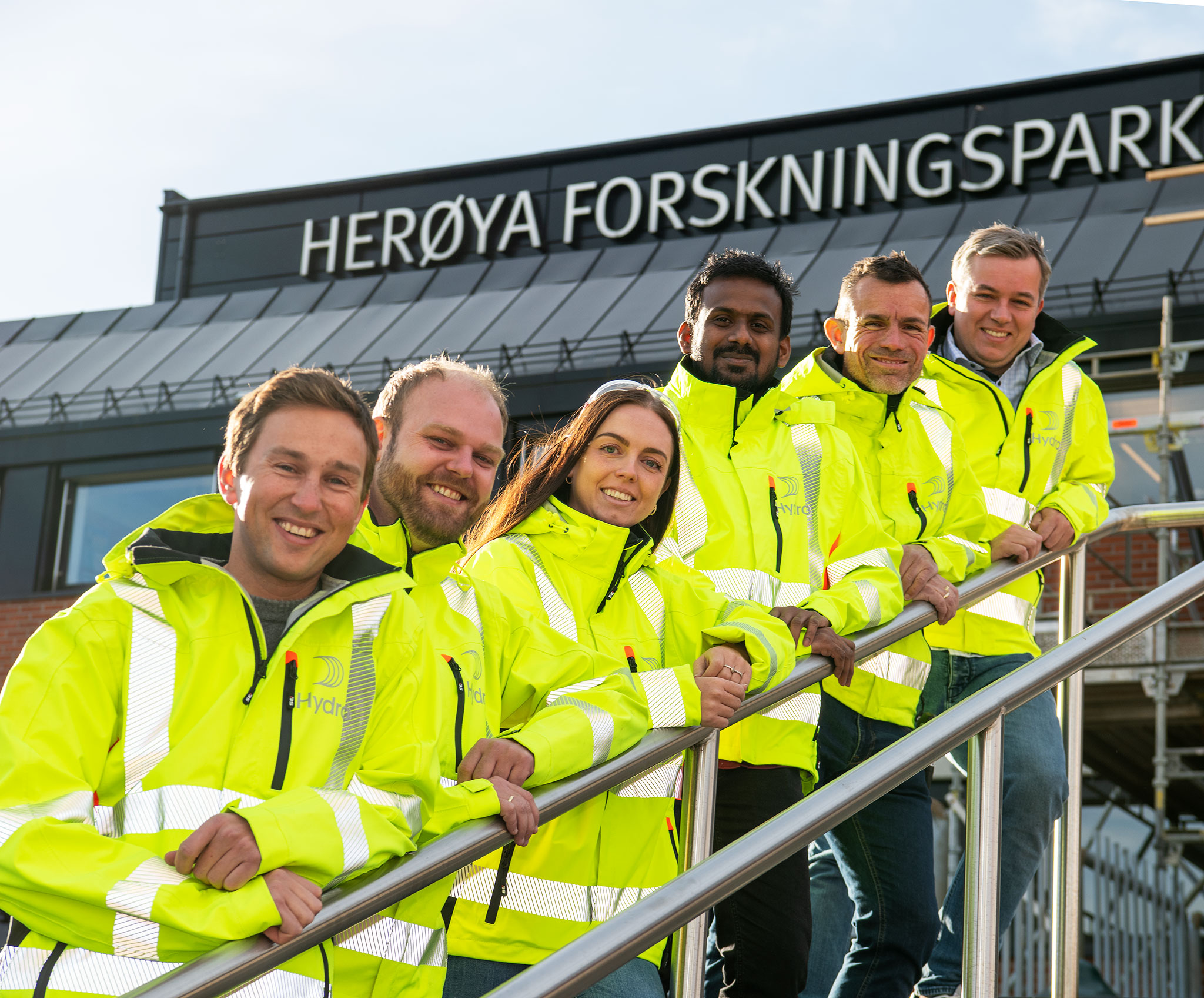 Six people, dressed in yellow jackets, pose, stand up a flight of stairs and lean against the railing outside a brick building. On the top of the building, large letters that read Herøya Forskningspark are partially displayed
