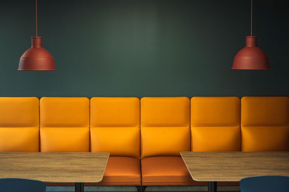 green wall, orange sofa, brown lamps down from ceiling, and two tables in the middle, part seating area in a canteen, artistic photo.