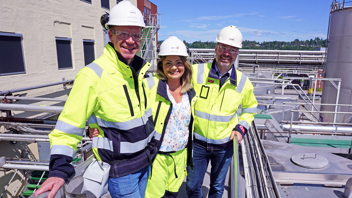 Three people, posing, wearing PPE, standing on top of steel tanks/process facility, blue sky