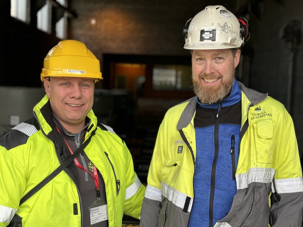 two men, posing, standing, smiling, wearing PPE, concret hall behind.