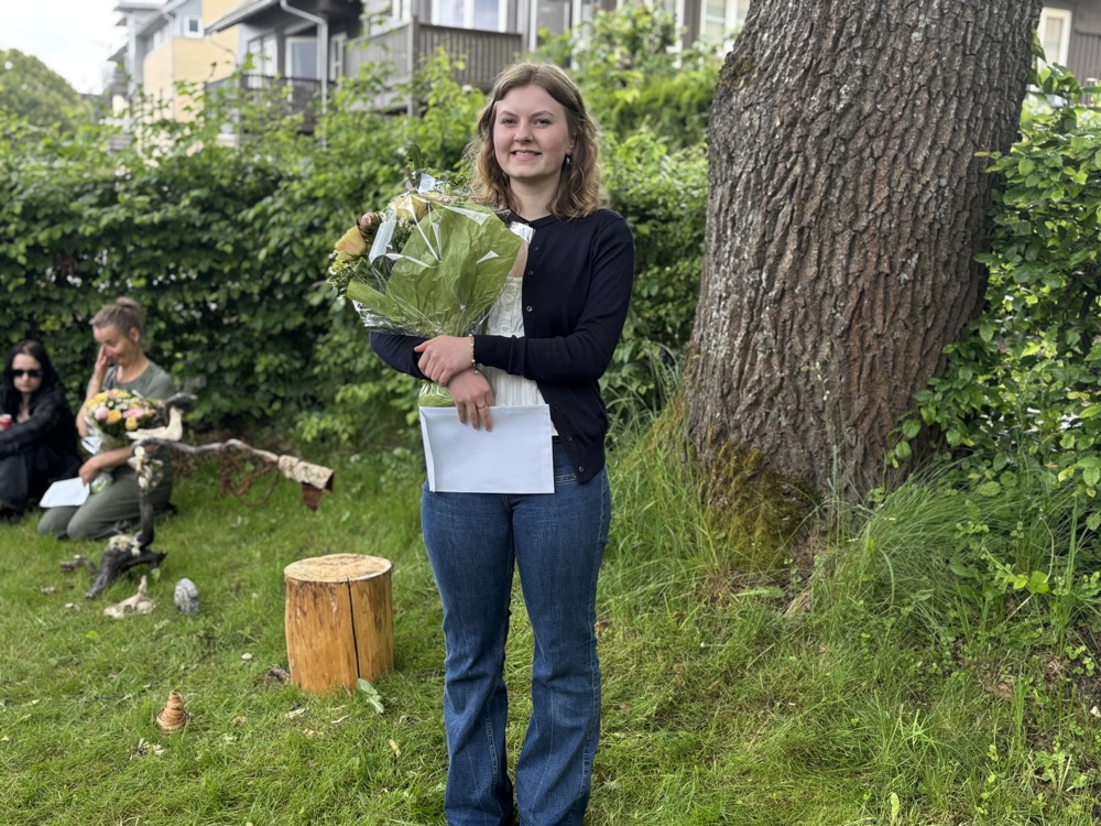 Ung kvinne med blomsterbukett og konvolutt i hendene, poserer, står ved en grønn hekk og stor trestamme.