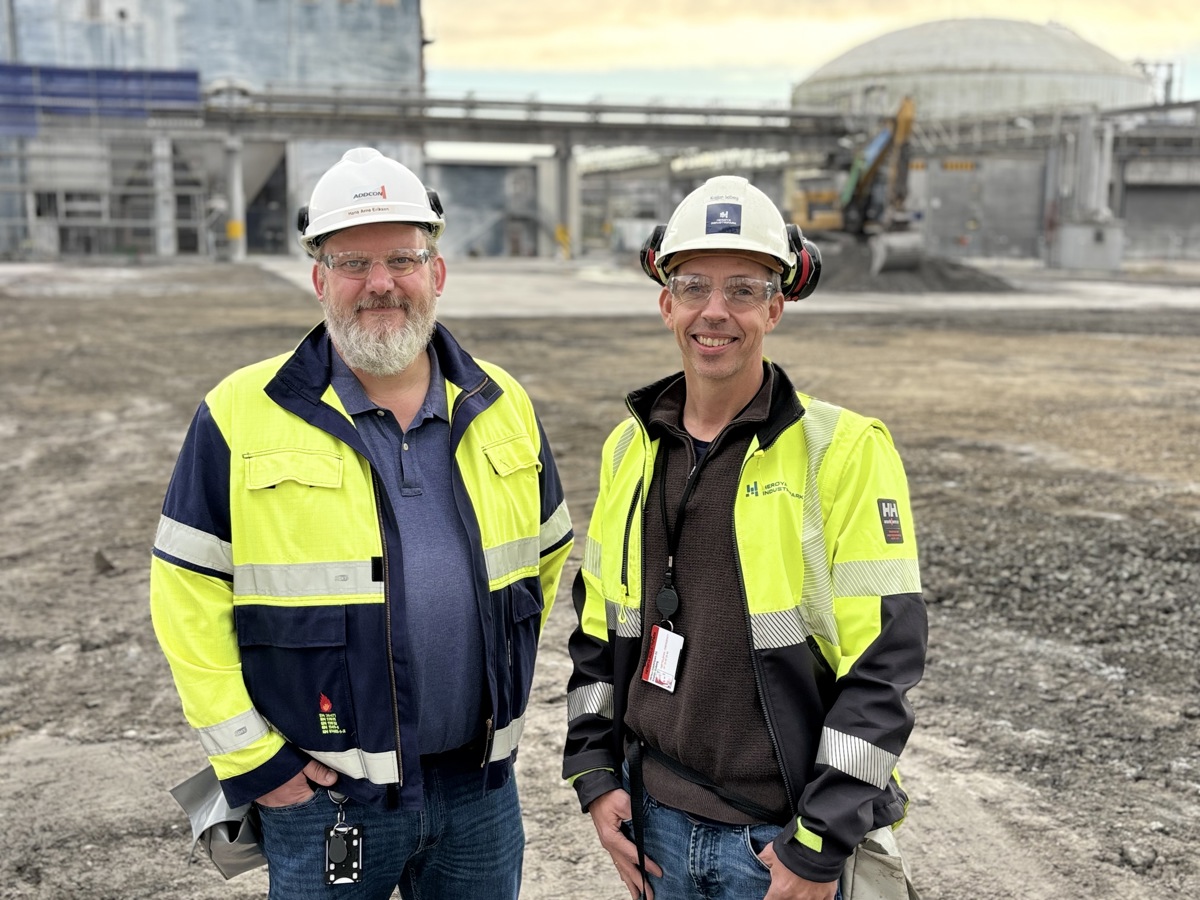 to menn, poserer, arbeidsklær, hvite hjelmer, industritomt i bakgrunnen