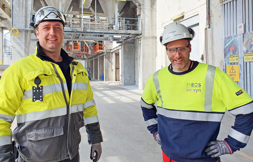 two men, wearing PPE, posing, outside production plant,