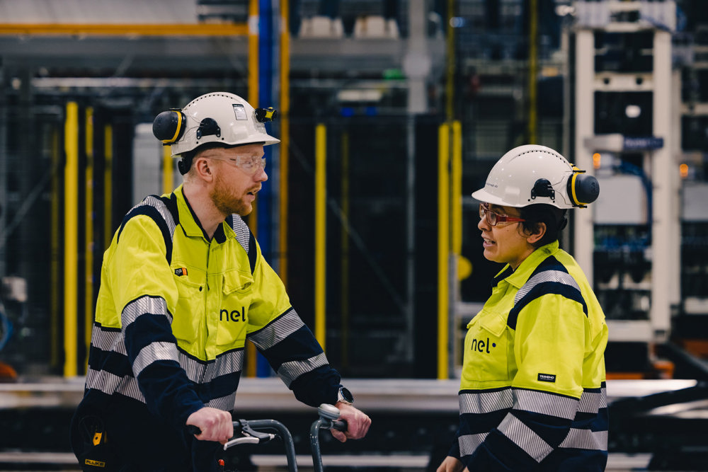 man and woman in dialog in production hall, both yellow jackets and white helmets