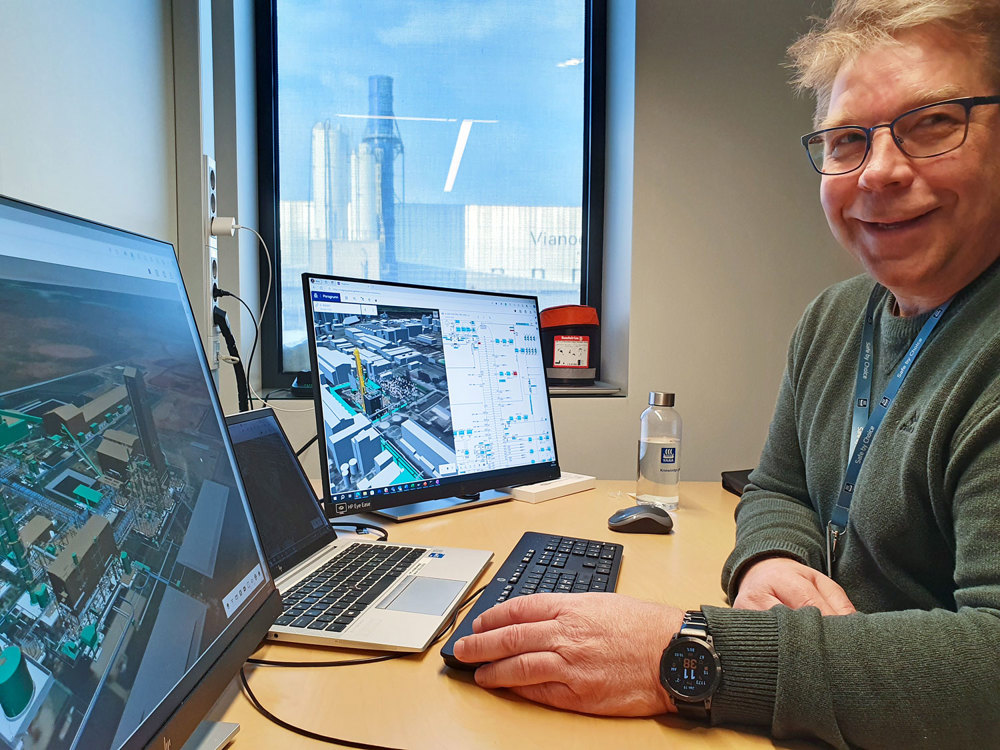 man with glasses sitting by his desk, computer and monitor in front of him, production plant outside his window