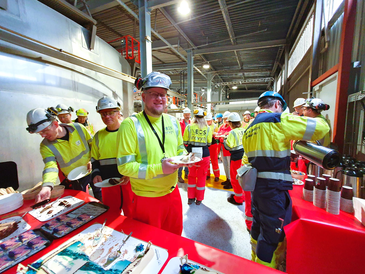 Kollegaer står i rekke for å forsyne seg med kake, markering, arbeidsantrekk røde bukser og gule gensere, hvite hjelmer, arrangement foregår i fabrikkhall.