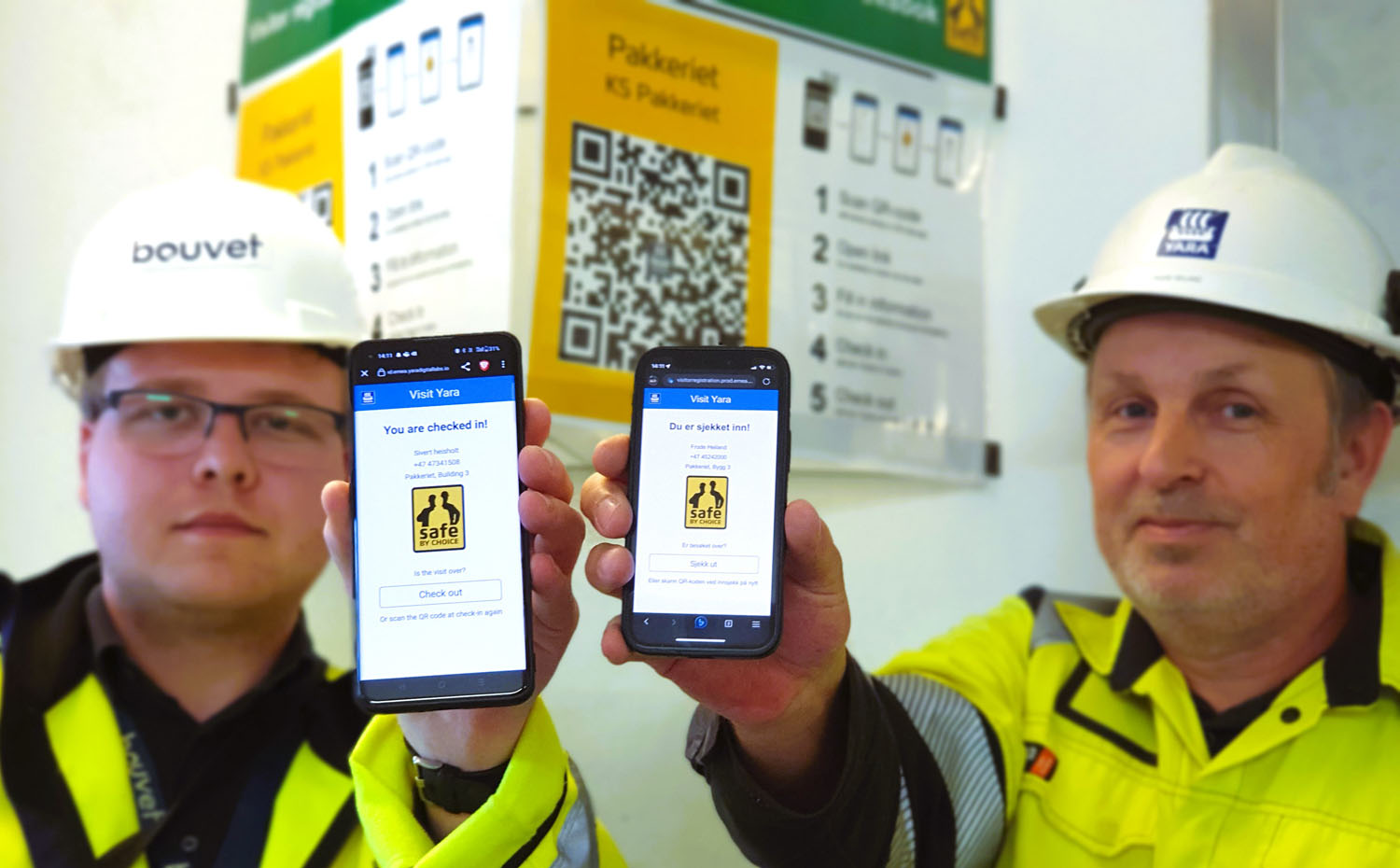 two men hold out their mobiles and show the visitor app. They wear yellow jackets and white helmets, office environment, user manual for app displayed on the wall behind.