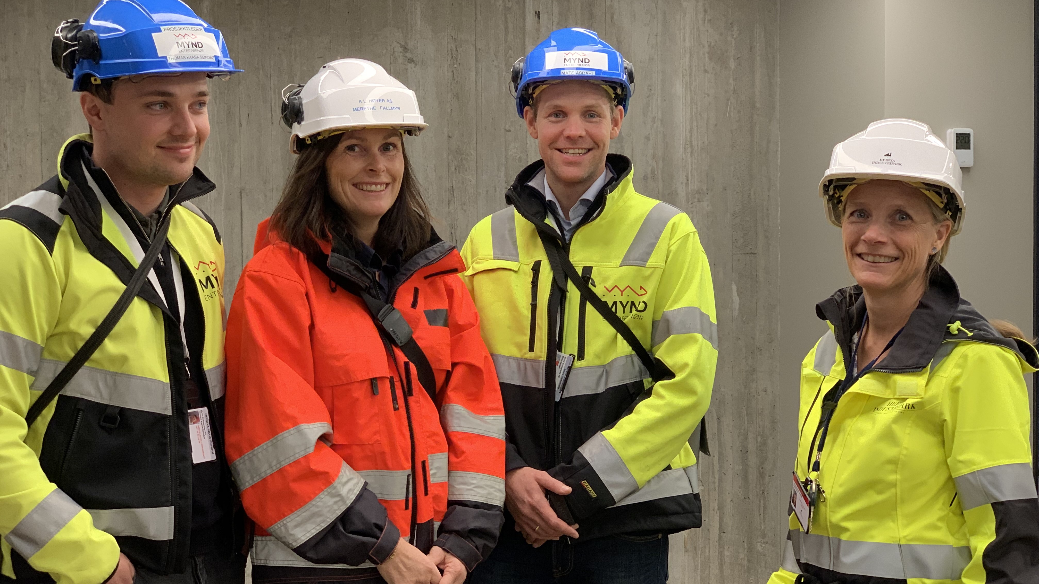 Four individuals in yellow and red work attire, blue and white helmets, posing, office environment, gray concrete in the background.