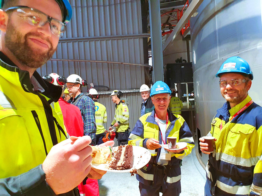 three men posing, wearing blue helmets and yellow jackets, eating cake and drinking coffee, standing in a production hall.